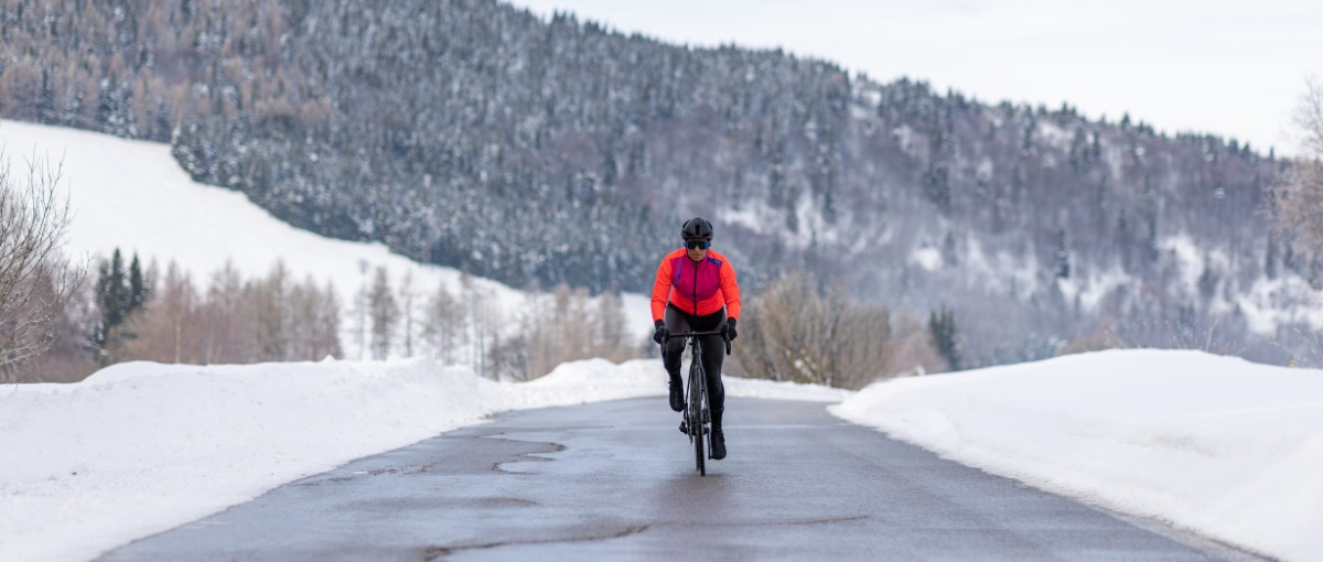 Cyclist in a fluro orange jacket riding on a snow-lined road in a mountainous area.
