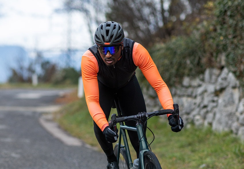 Cyclist wearing a helmet, sunglasses, and a sleeveless baselayer with fluro orange sleeves, riding on a road.