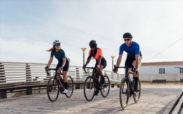 Three cyclists riding on a wooden boardwalk, wearing helmets and cycling apparel.