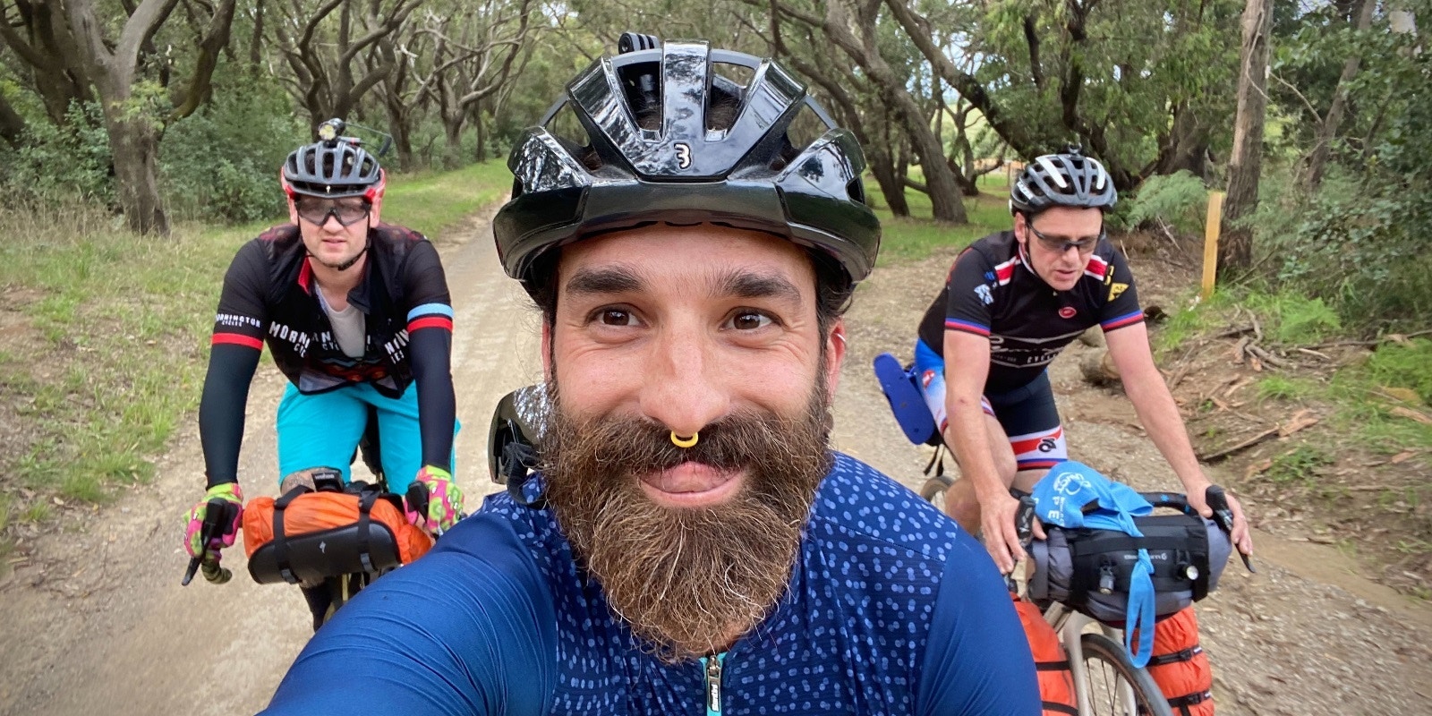 Three cyclists with loaded bikes riding on a forest trail, smiling at the camera.