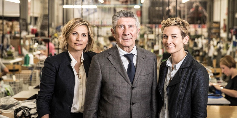 Three people, two women and one man, standing in a factory setting, dressed in business attire, with the man in the center wearing a suit and the women on either side.