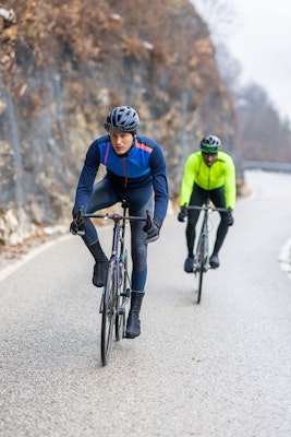 Two cyclists riding on a winding road, with one cyclist wearing a navy blue and red jacket and the other wearing a neon green jacket.
